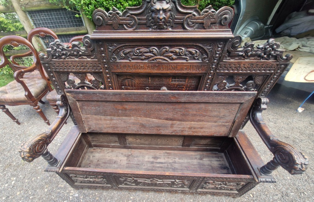 A late nineteenth-century dark carved oak hall bench featuring relief narrative carving, mask pediment, scroll arms, and a hinged seat. View XIX