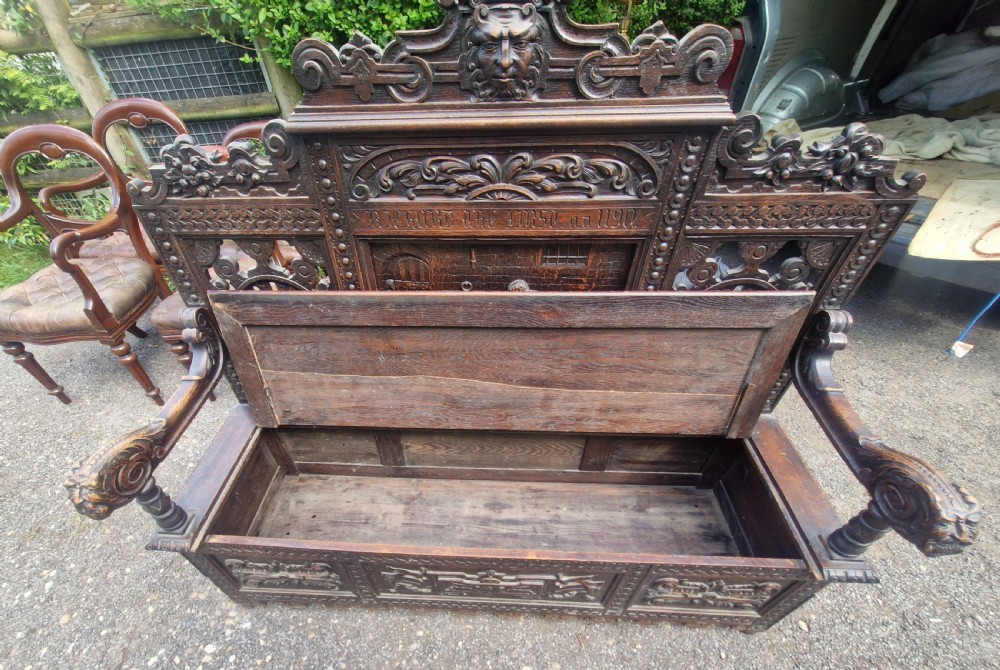 A late nineteenth-century dark carved oak hall bench featuring relief narrative carving, mask pediment, scroll arms, and a hinged seat. View XX