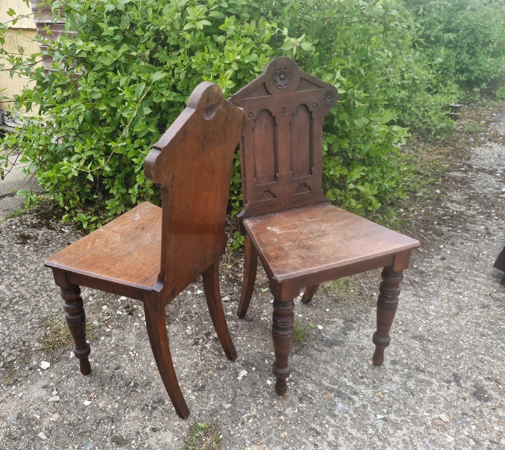 A matching pair of Victorian dark oak hall chairs featuring carved gothic arch backrests with flower rosettes, solid wood seats, and turned front legs. View II