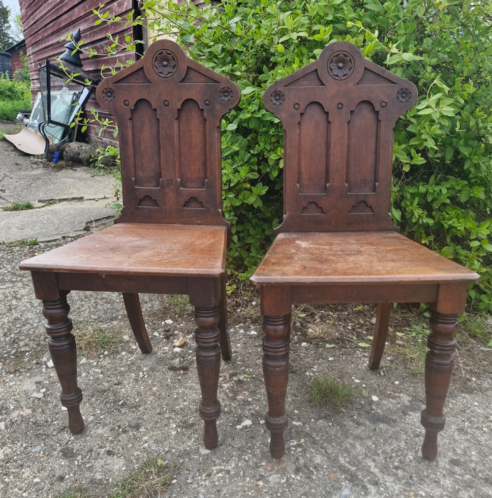 A matching pair of Victorian dark oak hall chairs featuring carved gothic arch backrests with flower rosettes, solid wood seats, and turned front legs. View IV