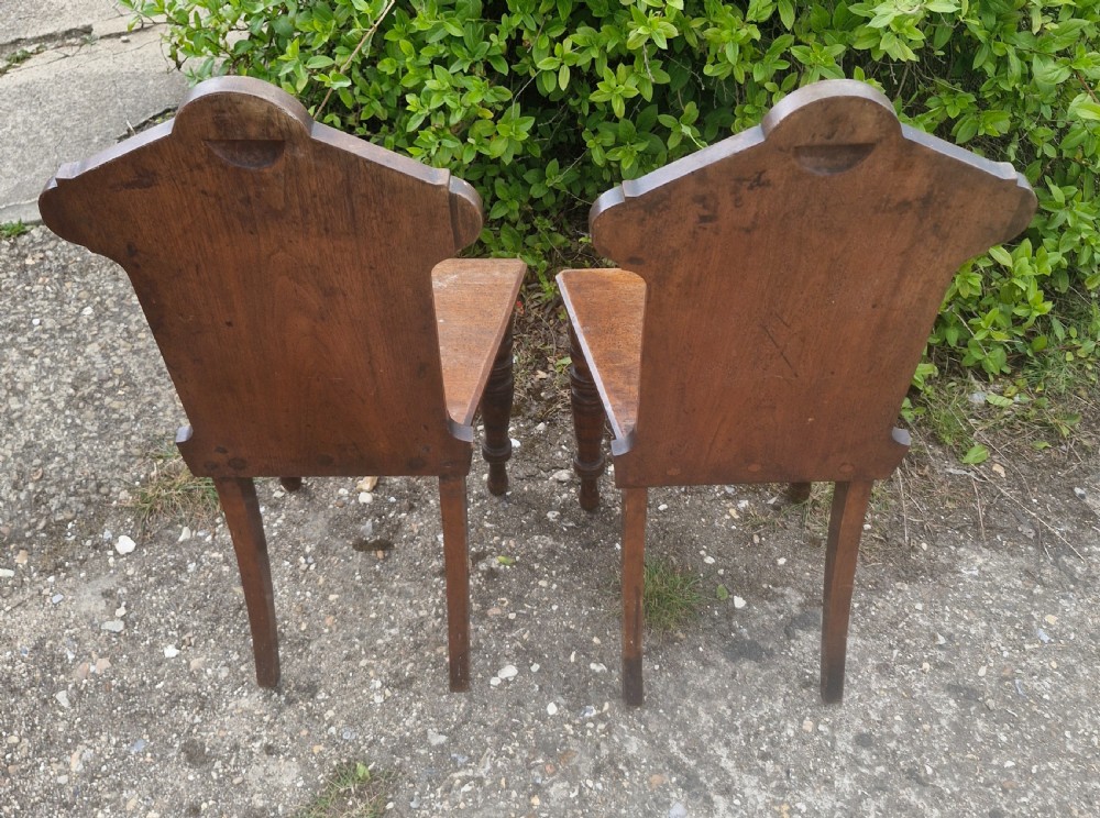 A matching pair of Victorian dark oak hall chairs featuring carved gothic arch backrests with flower rosettes, solid wood seats, and turned front legs. View V