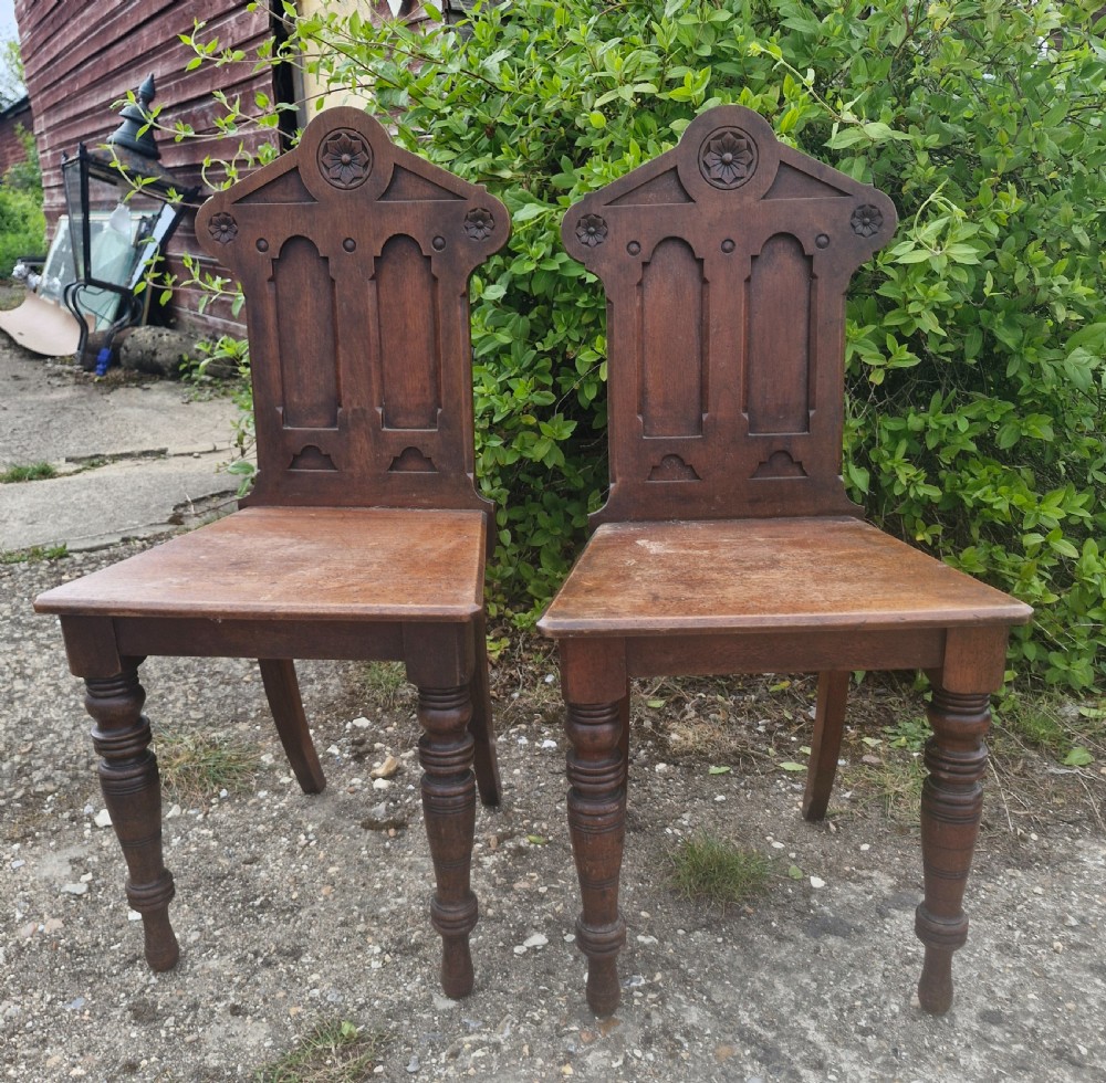 A matching pair of Victorian dark oak hall chairs featuring carved gothic arch backrests with flower rosettes, solid wood seats, and turned front legs. View VI