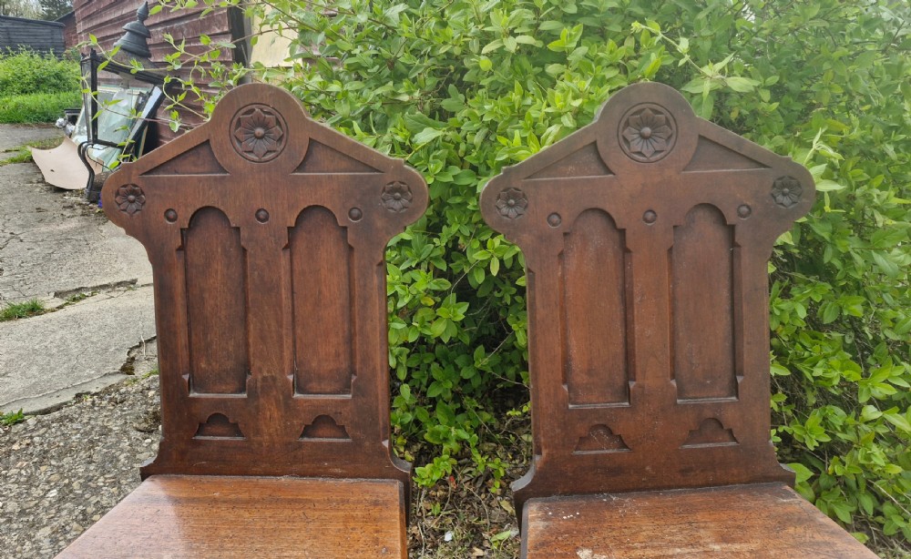 A matching pair of Victorian dark oak hall chairs featuring carved gothic arch backrests with flower rosettes, solid wood seats, and turned front legs. View VII
