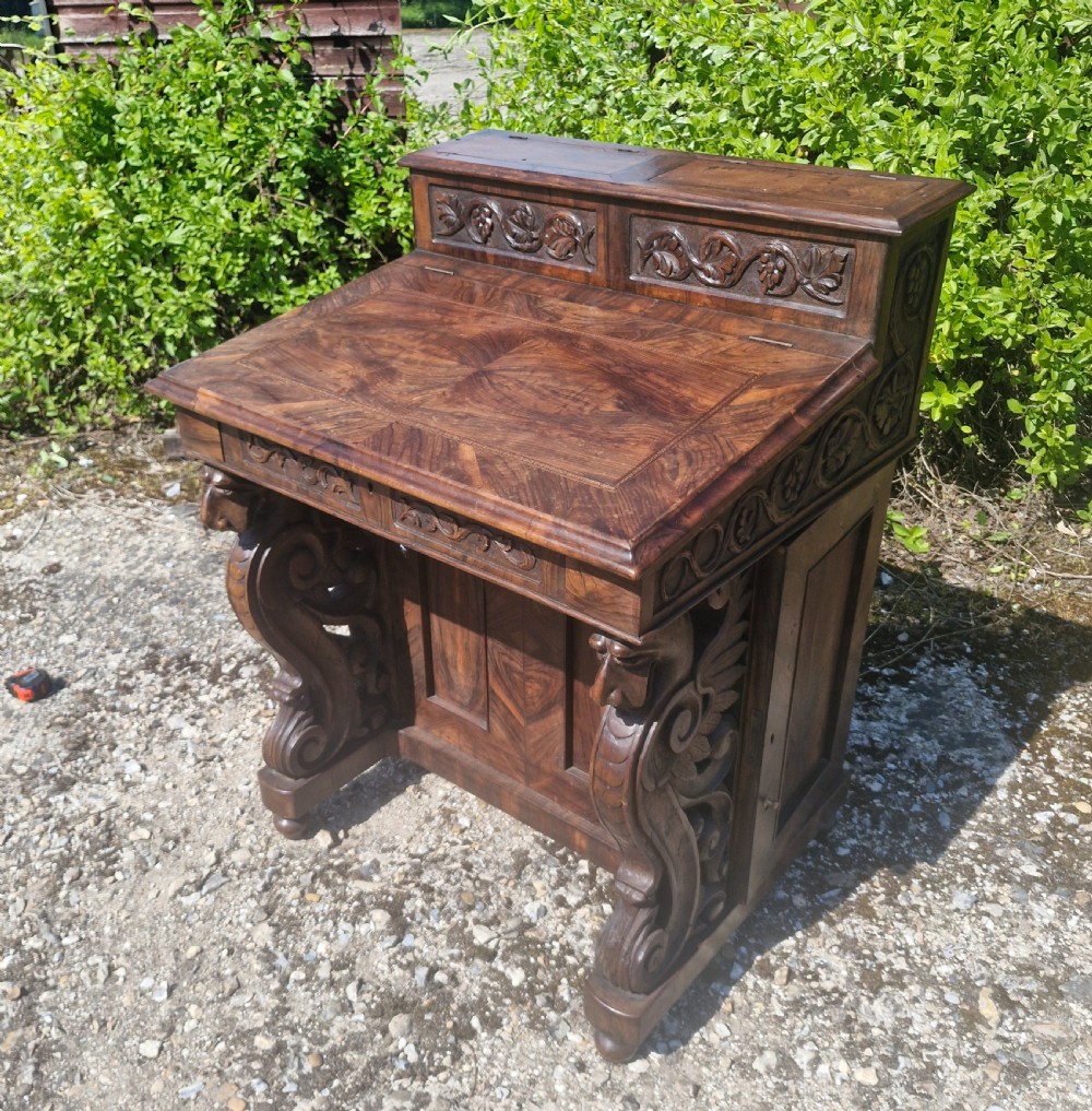 An antique carved olivewood Davenport writing desk with a sloped lid, foliage carvings, and decorative scrolled supports.