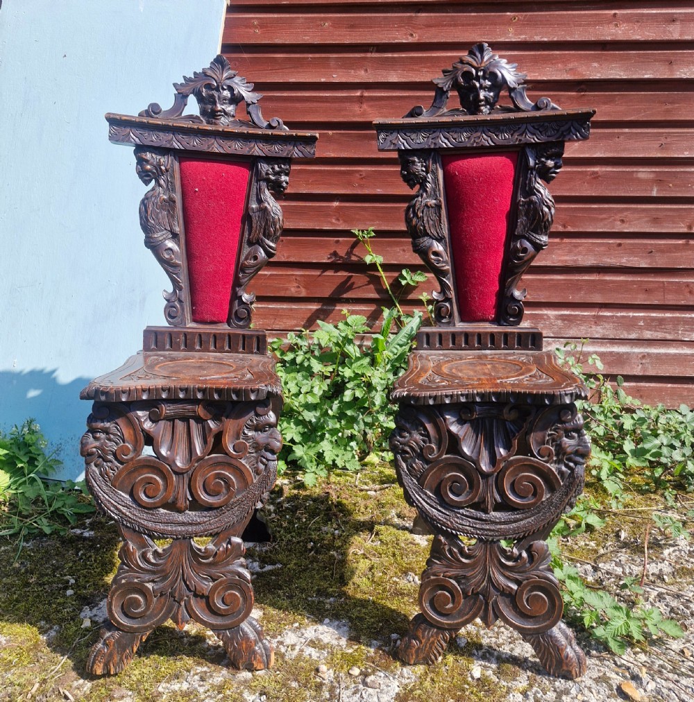 A pair of 19th-century carved walnut hall chairs featuring relief mascarons, scrollwork legs, and red velvet backrest panels.