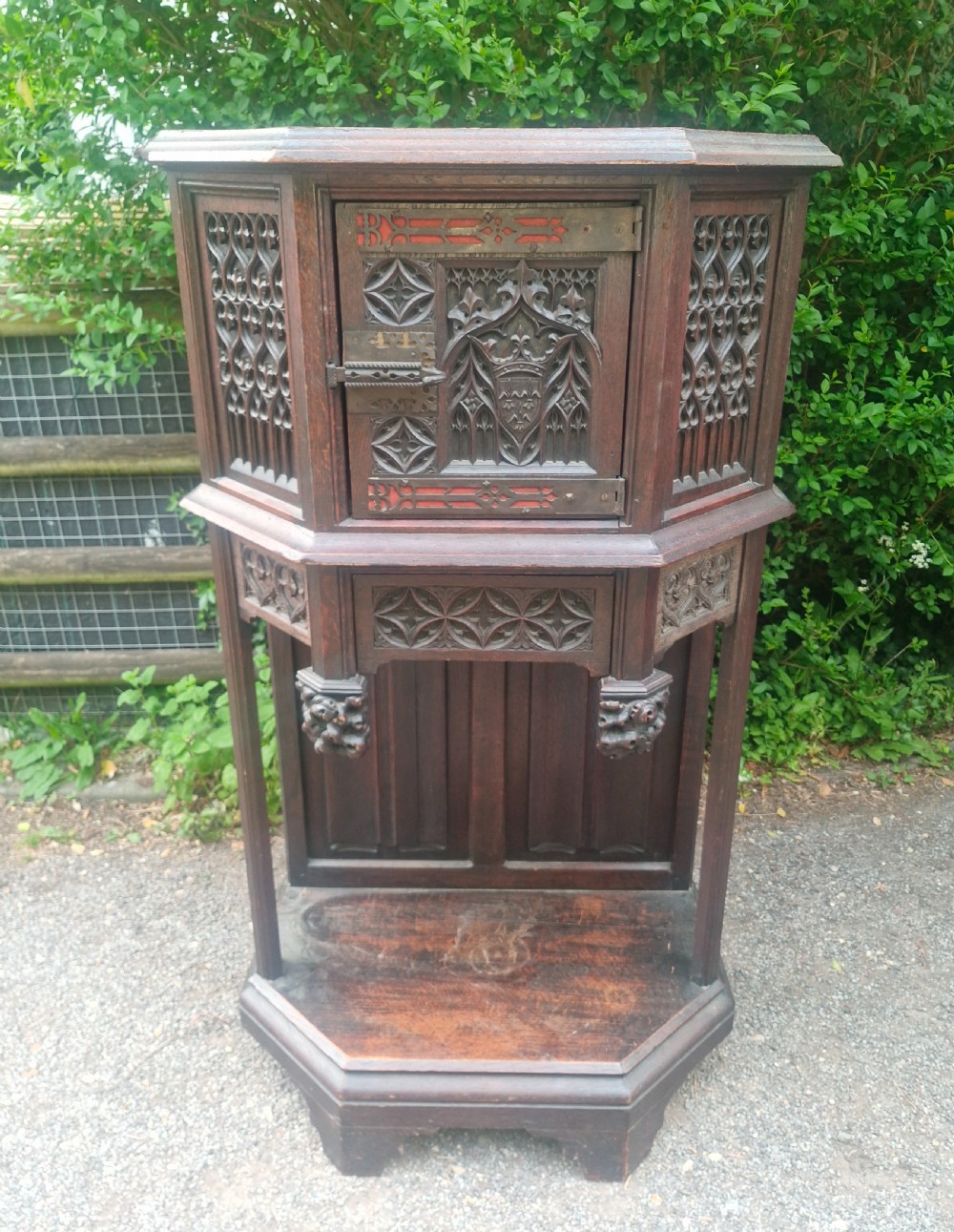 A Victorian Gothic Revival dark oak cabinet featuring intricately carved tracery panels, red metal accents, an armorial crest on the door, carved drop finials, and an open lower shelf.