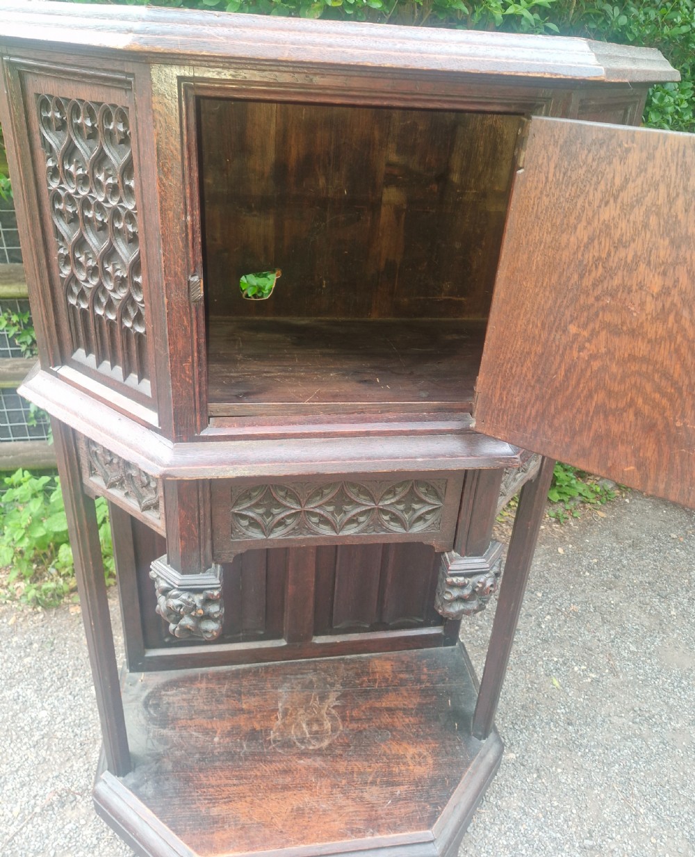A Victorian Gothic Revival dark oak cabinet featuring intricately carved tracery panels, red metal accents, an armorial crest on the door, carved drop finials, and an open lower shelf. View II