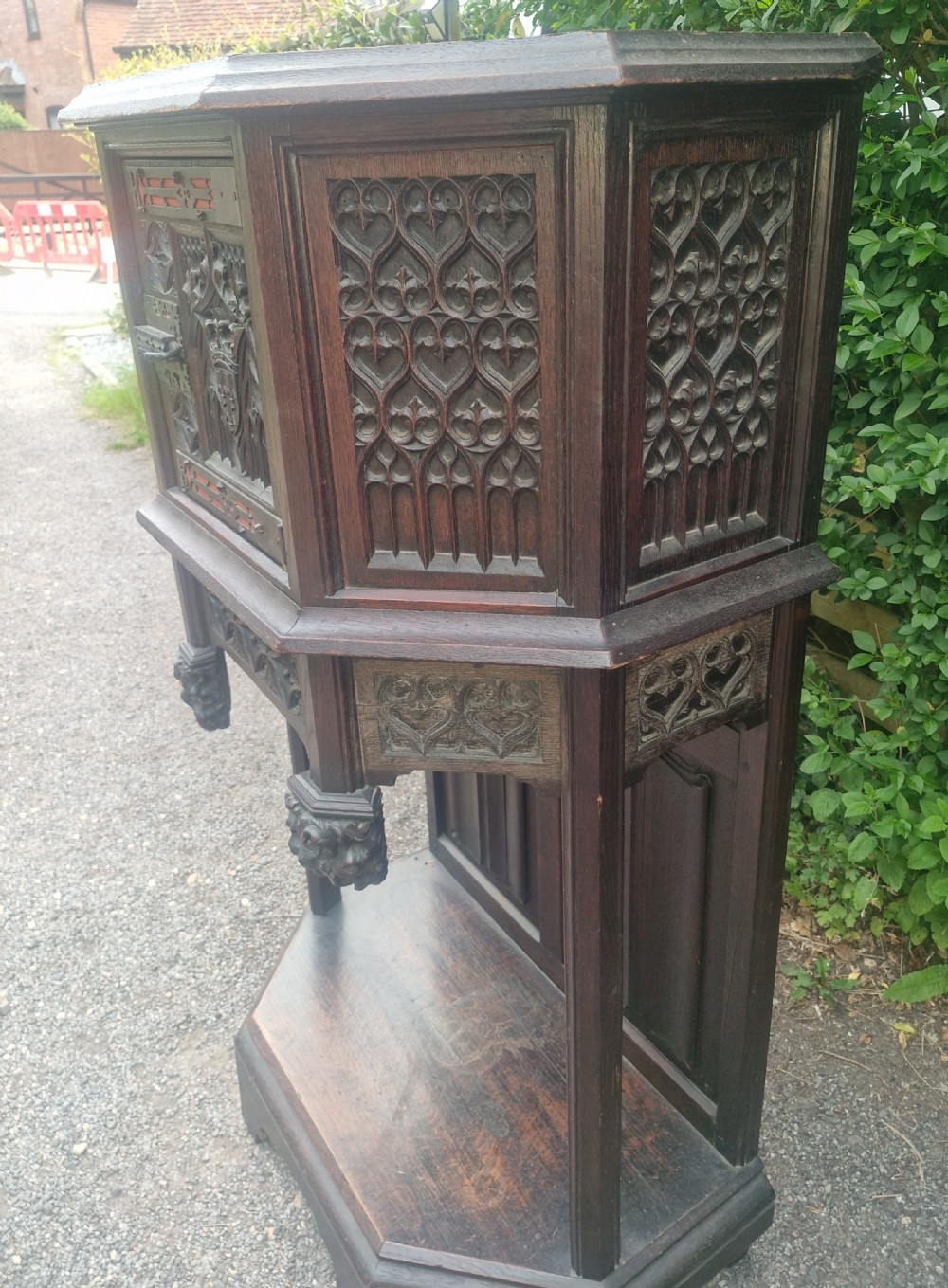 A Victorian Gothic Revival dark oak cabinet featuring intricately carved tracery panels, red metal accents, an armorial crest on the door, carved drop finials, and an open lower shelf. View III