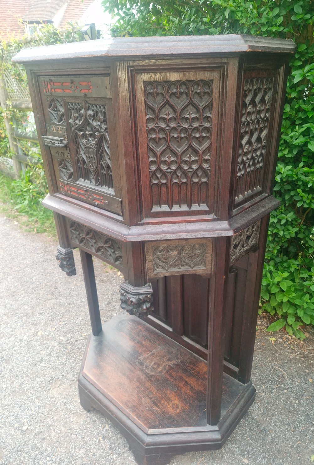 A Victorian Gothic Revival dark oak cabinet featuring intricately carved tracery panels, red metal accents, an armorial crest on the door, carved drop finials, and an open lower shelf. View IV