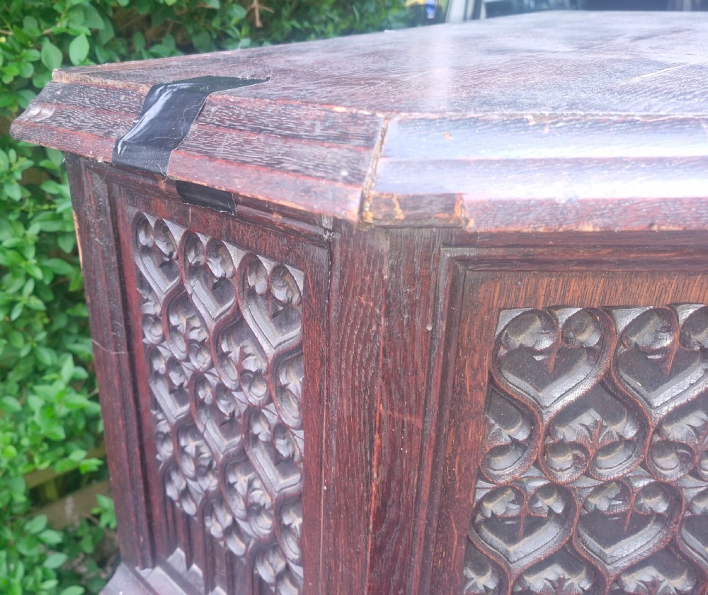 A Victorian Gothic Revival dark oak cabinet featuring intricately carved tracery panels, red metal accents, an armorial crest on the door, carved drop finials, and an open lower shelf. View V