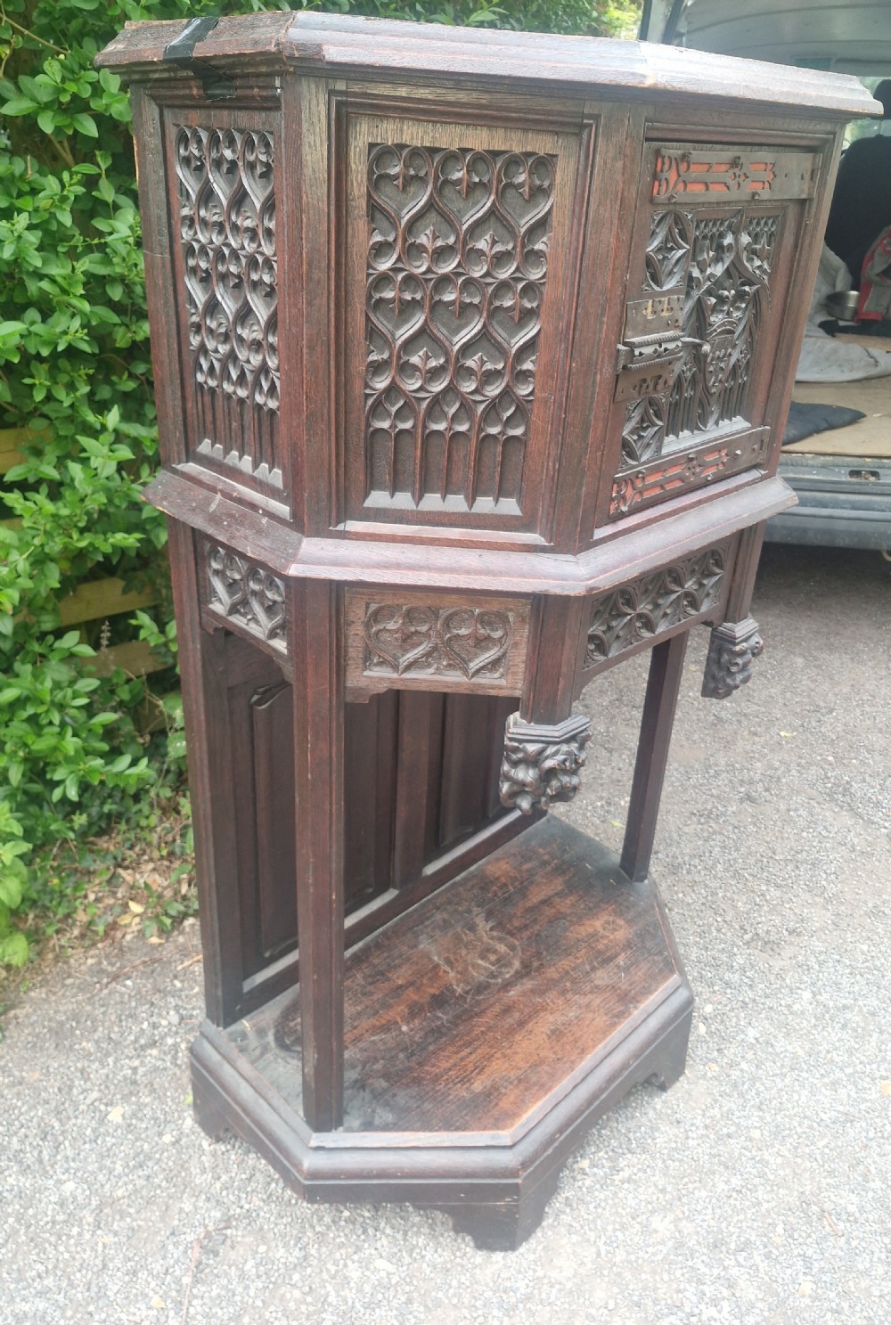 A Victorian Gothic Revival dark oak cabinet featuring intricately carved tracery panels, red metal accents, an armorial crest on the door, carved drop finials, and an open lower shelf. View VI
