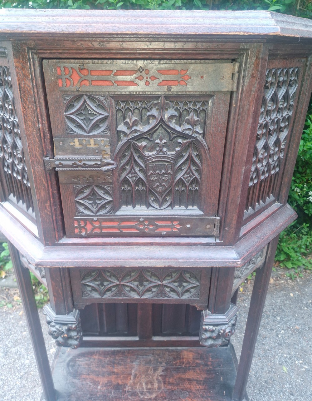 A Victorian Gothic Revival dark oak cabinet featuring intricately carved tracery panels, red metal accents, an armorial crest on the door, carved drop finials, and an open lower shelf. View VII