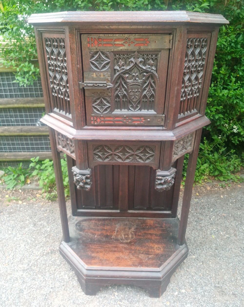 A Victorian Gothic Revival dark oak cabinet featuring intricately carved tracery panels, red metal accents, an armorial crest on the door, carved drop finials, and an open lower shelf. View VIII