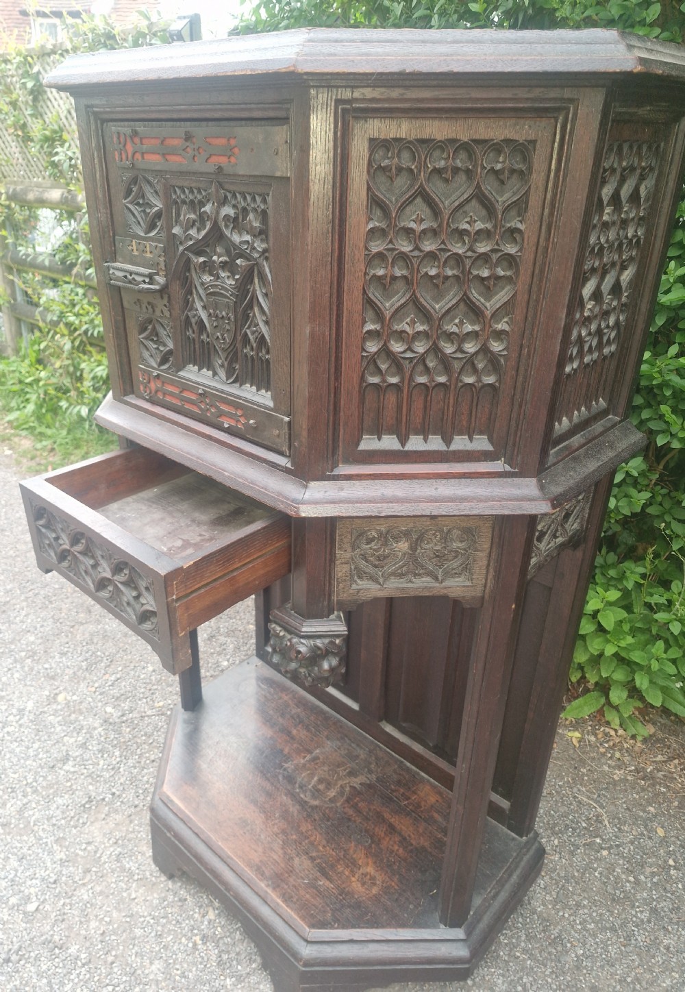 A Victorian Gothic Revival dark oak cabinet featuring intricately carved tracery panels, red metal accents, an armorial crest on the door, carved drop finials, and an open lower shelf. View IX