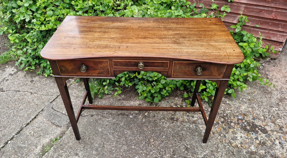 An antique Regency mahogany side table featuring a curved front apron, three inlaid drawers with turned brass handles, and square tapered legs connected by stretchers.