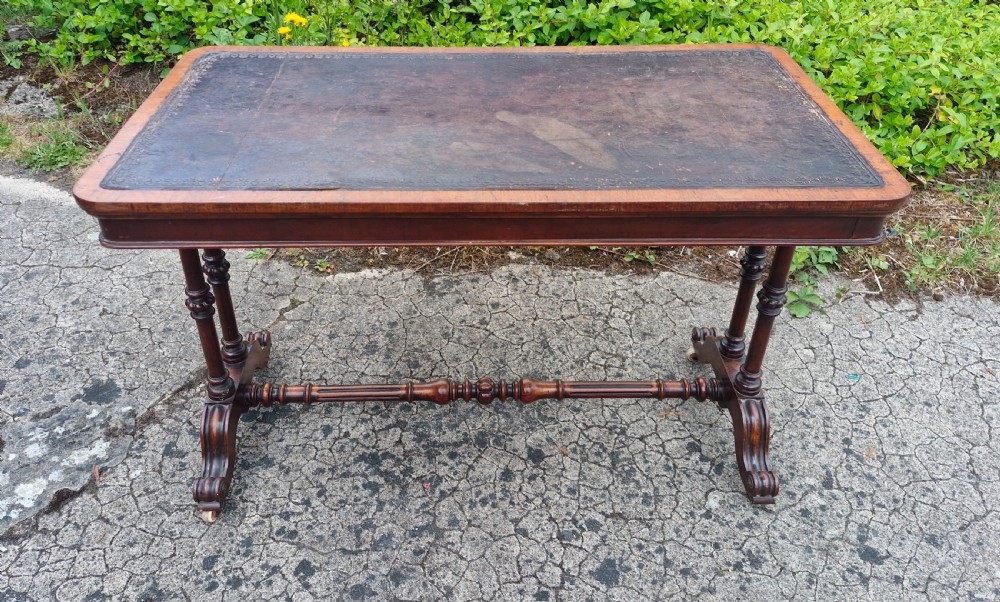 A mid-Victorian rectangular walnut library table featuring an inset dark leather top, turned dual end supports, a central turned stretcher, and carved legs with castors.