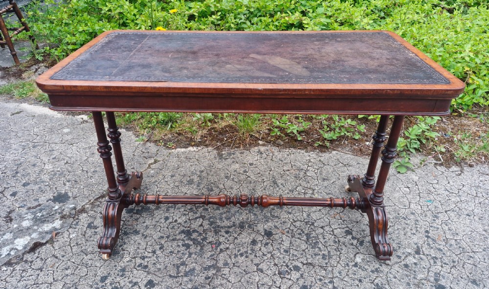 A mid-Victorian rectangular walnut library table featuring an inset dark leather top, turned dual end supports, a central turned stretcher, and carved legs with castors. View V