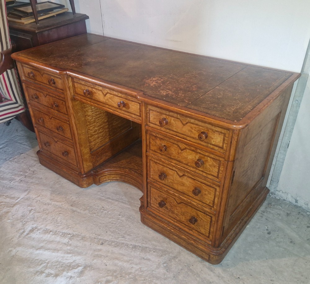 A Victorian Hungarian ash pedestal writing desk featuring a worn leather top, turned wooden drawer knobs, and inlaid geometric crossbanding.