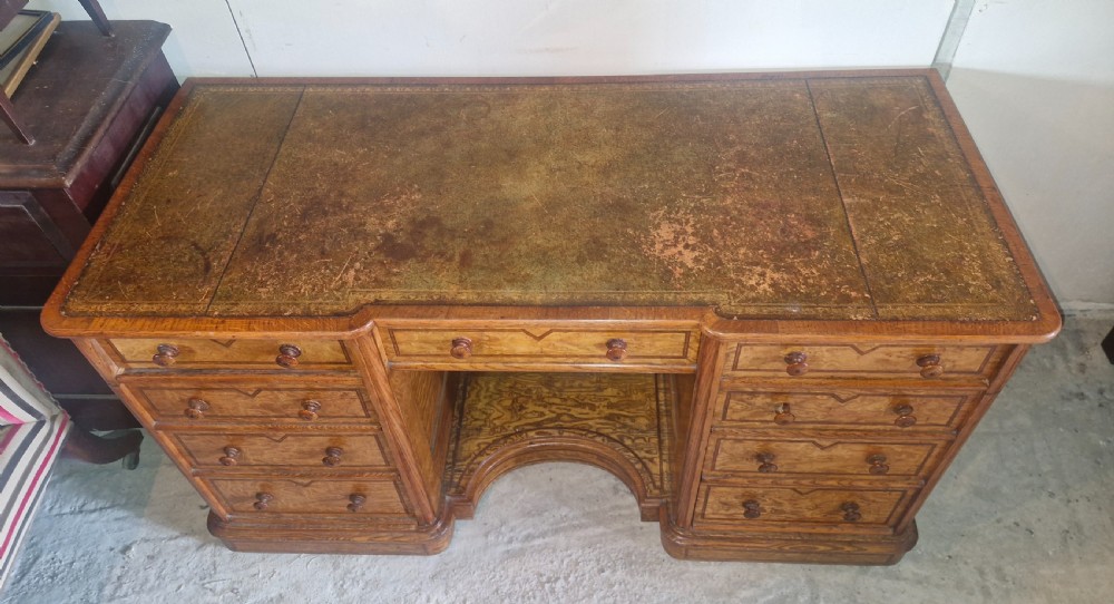 A Victorian Hungarian ash pedestal writing desk featuring a worn leather top, turned wooden drawer knobs, and inlaid geometric crossbanding. View VI
