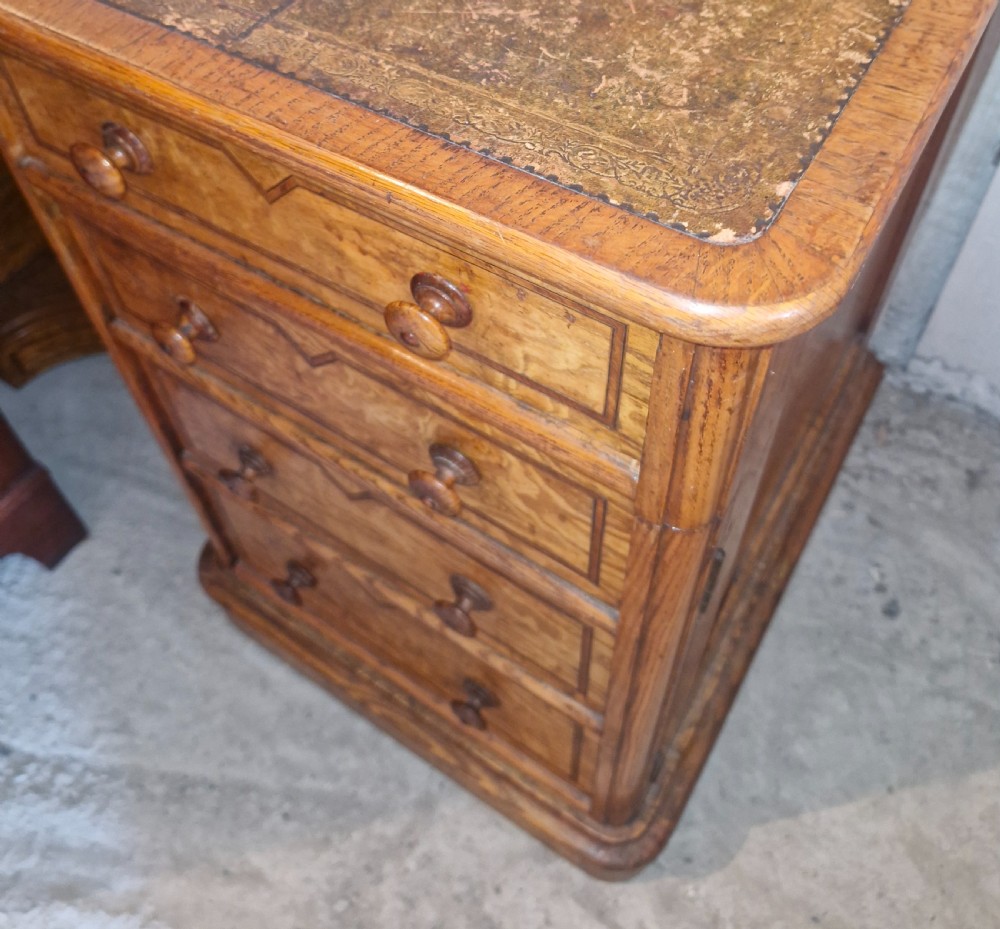A Victorian Hungarian ash pedestal writing desk featuring a worn leather top, turned wooden drawer knobs, and inlaid geometric crossbanding. View X