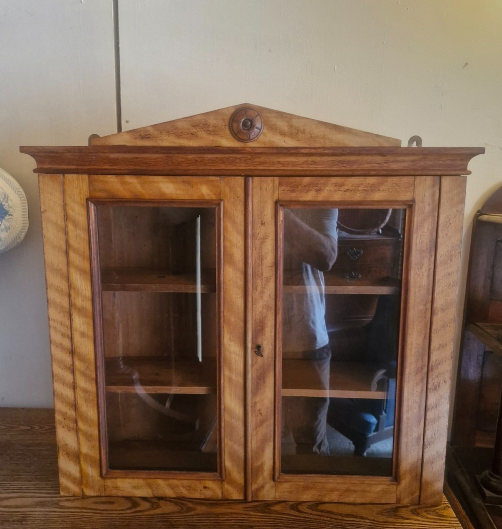 A Mid-Victorian satinbirch wall cabinet featuring two glazed doors, internal wooden shelves, and a pediment top with a central turned round patera.