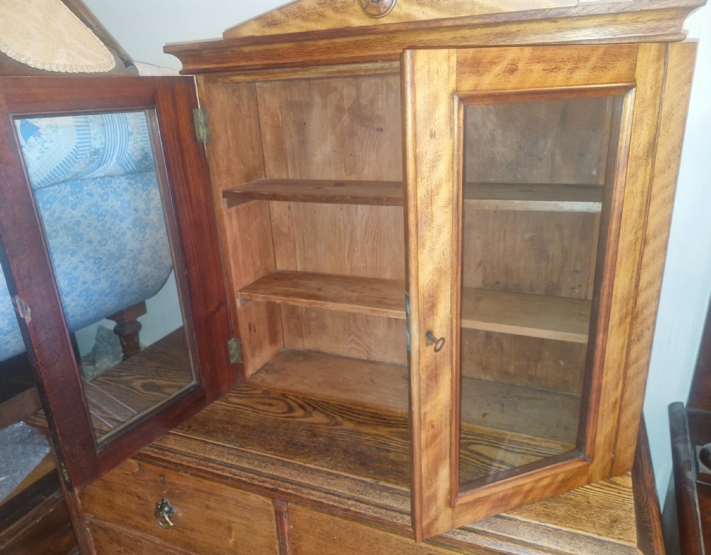 A Mid-Victorian satinbirch wall cabinet featuring two glazed doors, internal wooden shelves, and a pediment top with a central turned round patera. View II