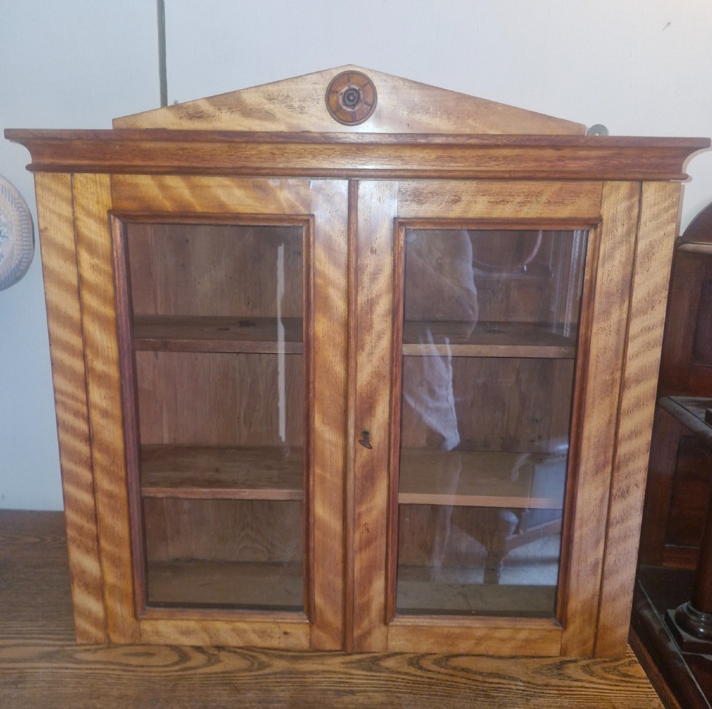 A Mid-Victorian satinbirch wall cabinet featuring two glazed doors, internal wooden shelves, and a pediment top with a central turned round patera. View III