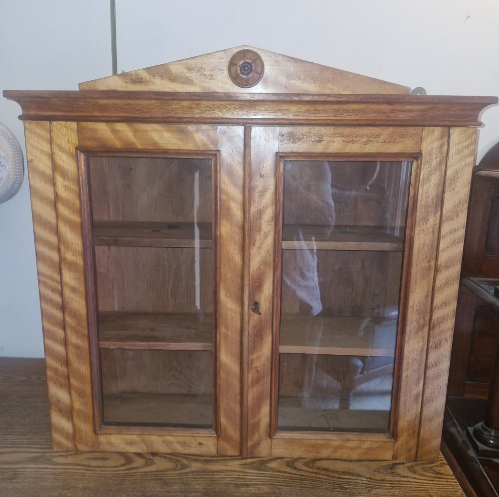 A Mid-Victorian satinbirch wall cabinet featuring two glazed doors, internal wooden shelves, and a pediment top with a central turned round patera. View VI