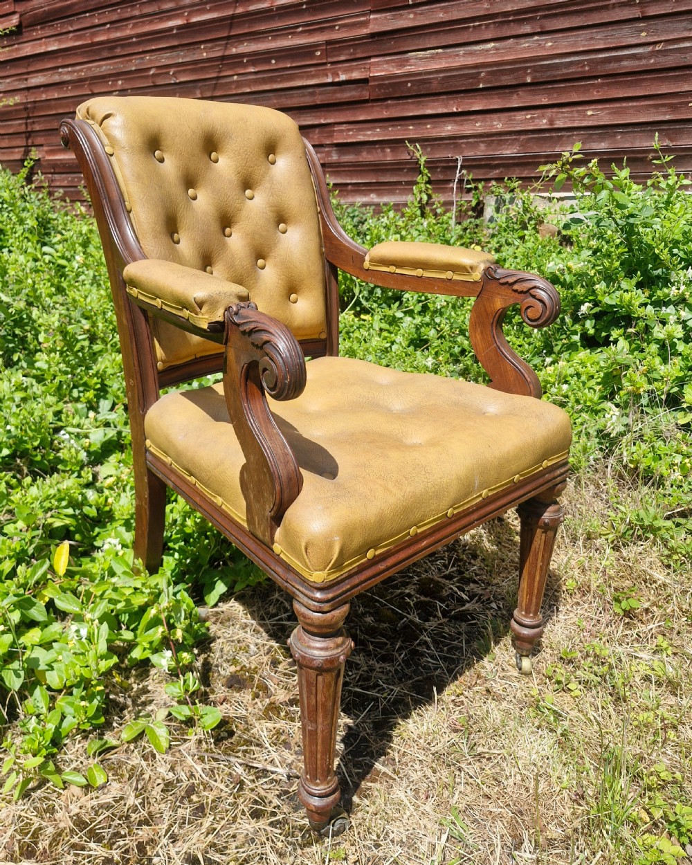 A mid-19th century mahogany desk armchair with tan button-tufted upholstery, scroll carved armrests, fluted front legs, and castors. View III
