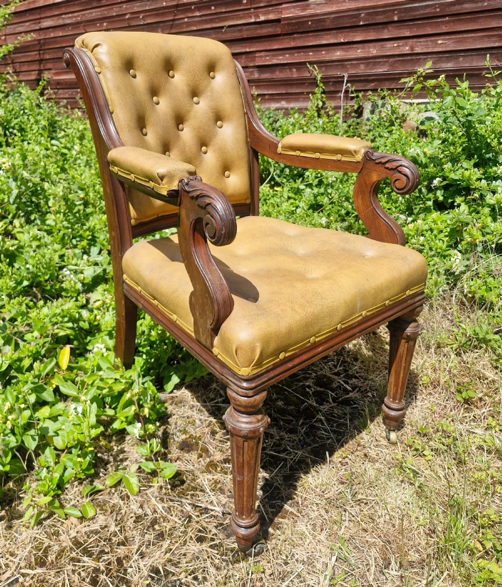 A mid-19th century mahogany desk armchair with tan button-tufted upholstery, scroll carved armrests, fluted front legs, and castors. View V