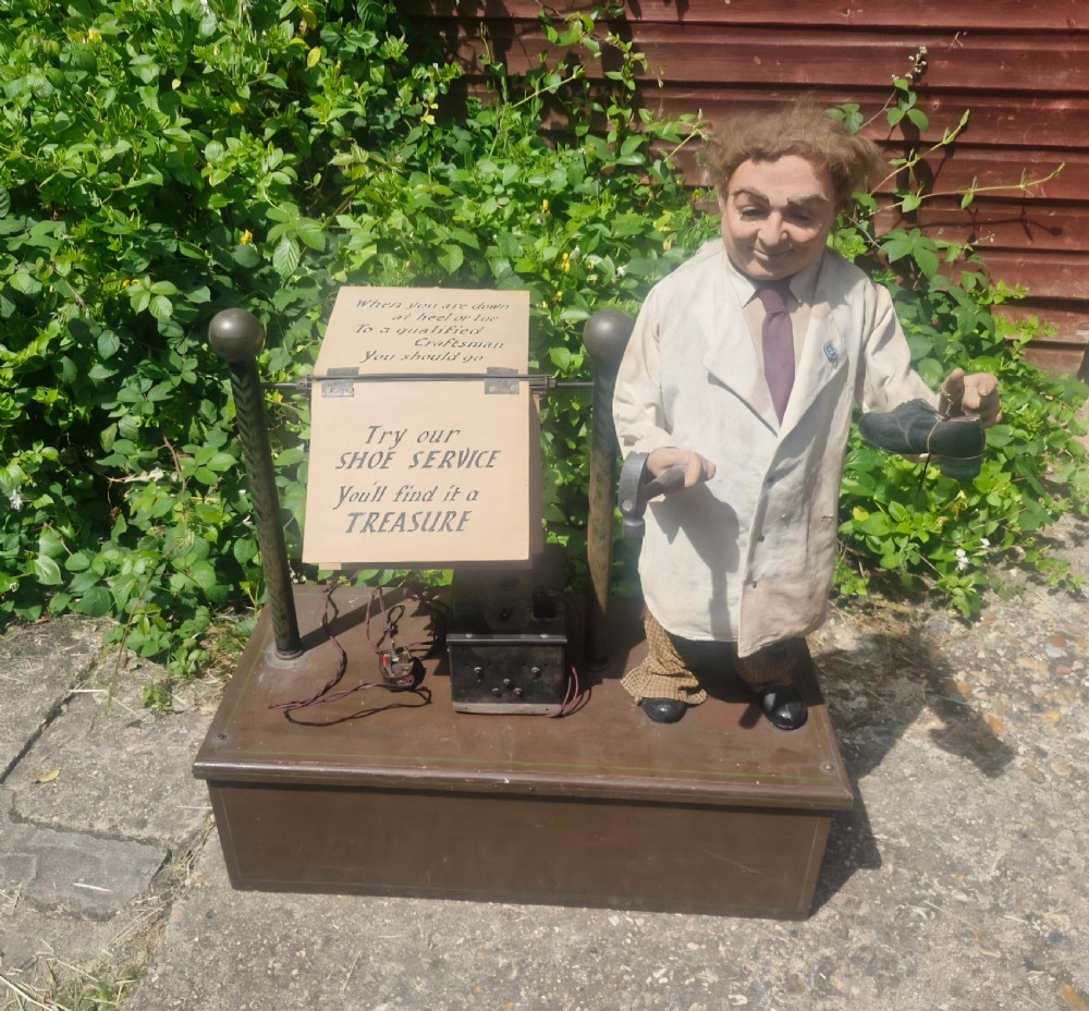An automated cobbler shop display model featuring a mechanised figure in a lab coat holding a shoe beside a mechanical signage mechanism on a dark wooden plinth.