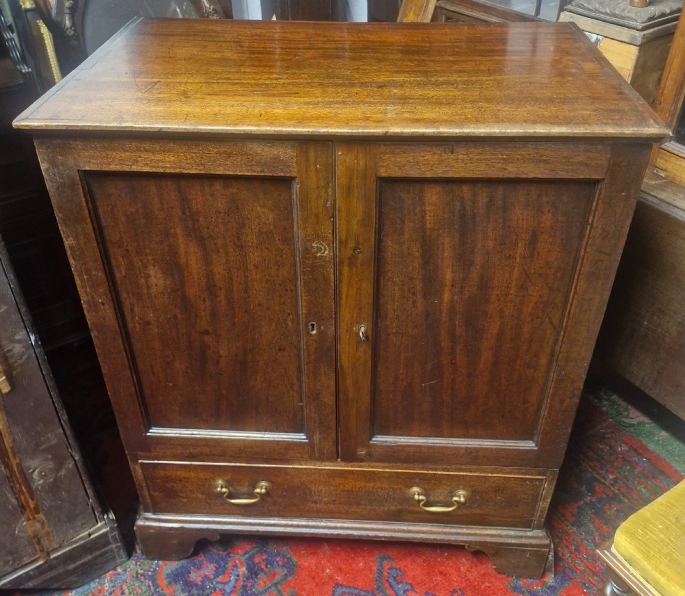 A late Georgian mahogany cupboard with two panelled doors positioned over a single wide drawer with twin brass handles, resting on bracket feet.