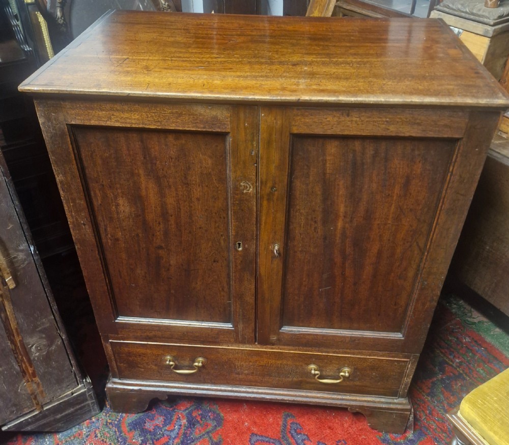 A late Georgian mahogany cupboard with two panelled doors positioned over a single wide drawer with twin brass handles, resting on bracket feet. View V