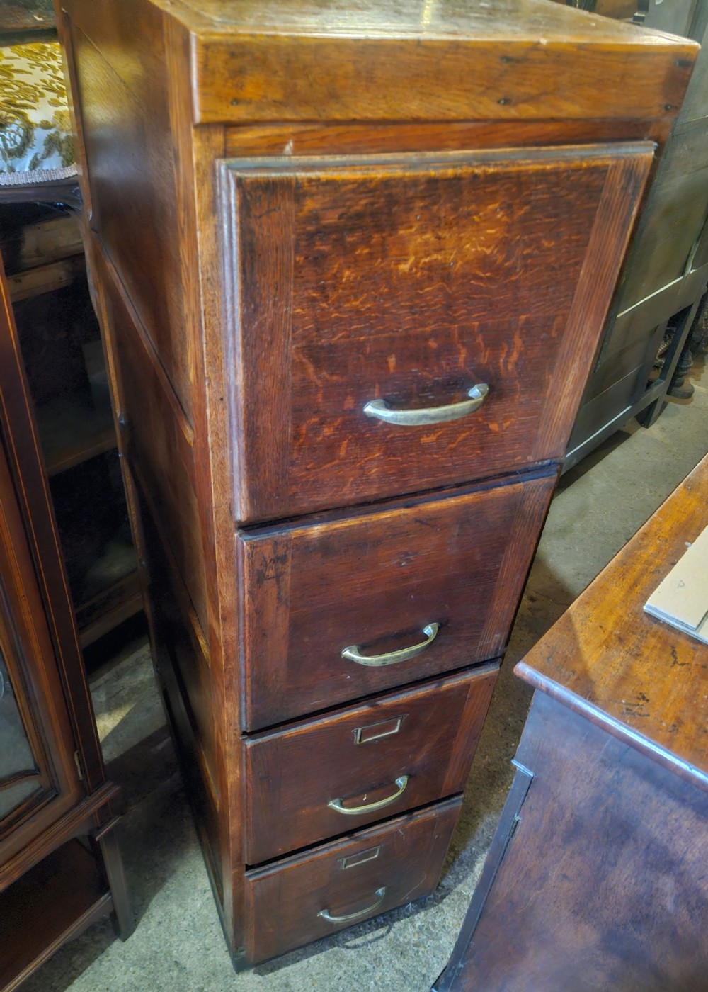 A tall oak four-drawer vertical filing cabinet featuring panelled sides and brass cup handles. View IV