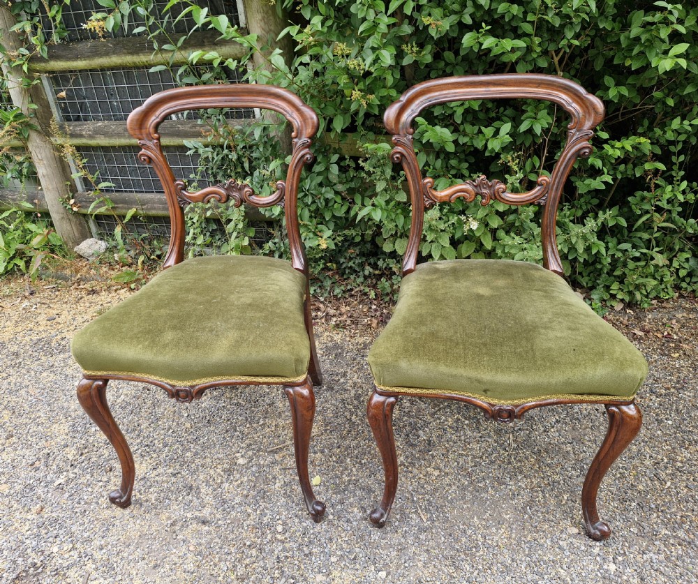 A pair of early Victorian rosewood balloon back side chairs featuring carved foliate splats, cabriole front legs, and green velvet upholstered seats.