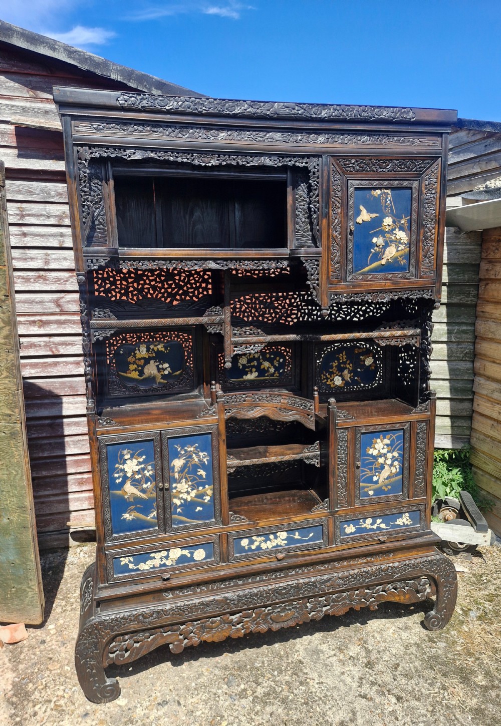 An intricately carved dark wood Japanese cabinet featuring staggered open shelves, small cupboards, and blue panels detailed with raised floral and bird inlays.