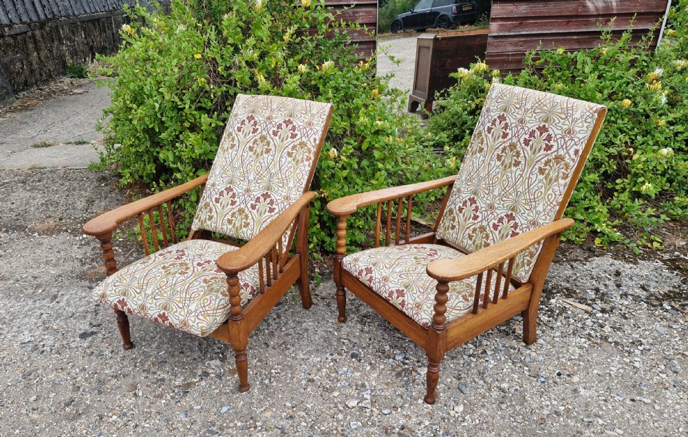 A pair of light oak Arts and Crafts reclining armchairs featuring turned front legs, spindled arm supports, and botanical pattern upholstery.