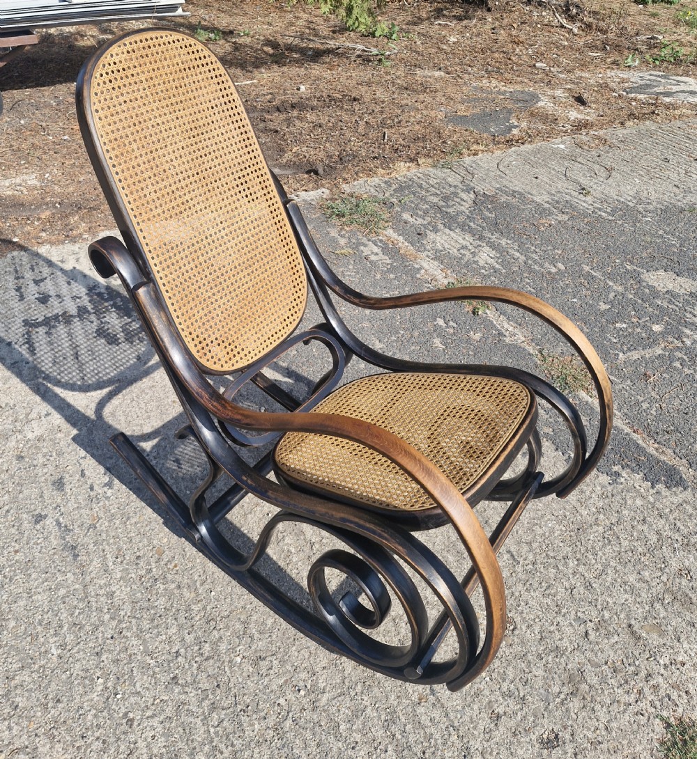 A dark brown bentwood rocking armchair featuring prominent scrollwork side frames with woven rattan cane backrest and seat panels. View IV