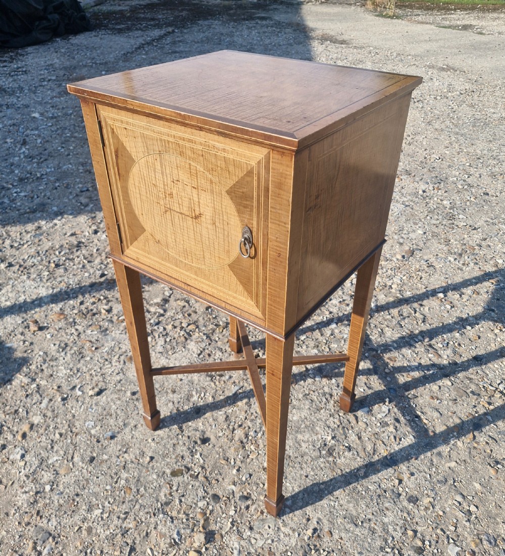 An Edwardian satinwood bedside cabinet featuring fine line stringing, an oval inlay design on the single door, square tapered legs, and an X-frame stretcher.