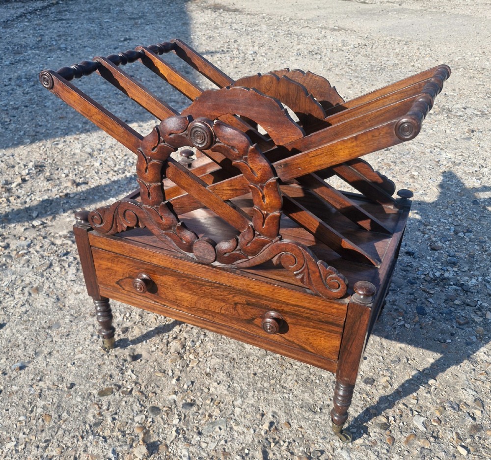 A Regency rosewood Canterbury featuring slatted division compartments, central carved leaf wreath motifs, a base drawer with turned handles, and turned legs on brass castors.