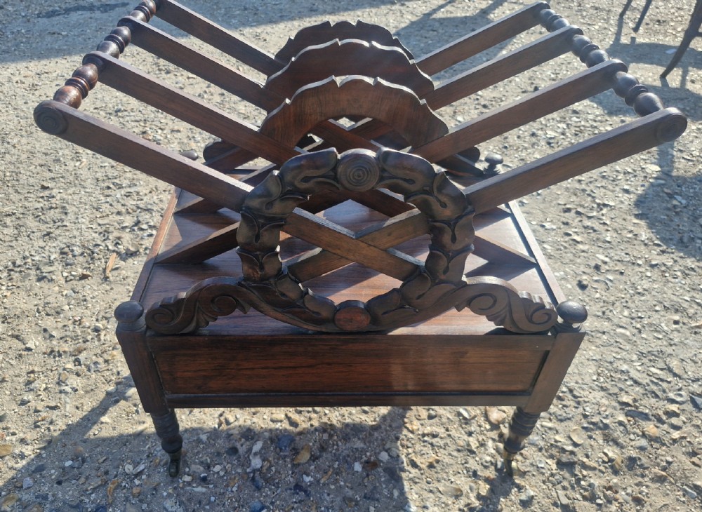 A Regency rosewood Canterbury featuring slatted division compartments, central carved leaf wreath motifs, a base drawer with turned handles, and turned legs on brass castors. View II