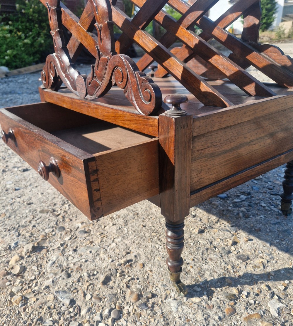 A Regency rosewood Canterbury featuring slatted division compartments, central carved leaf wreath motifs, a base drawer with turned handles, and turned legs on brass castors. View III