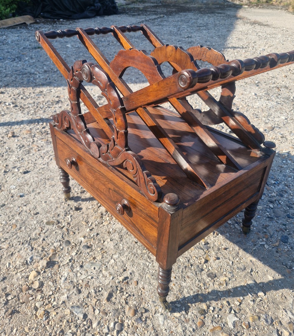 A Regency rosewood Canterbury featuring slatted division compartments, central carved leaf wreath motifs, a base drawer with turned handles, and turned legs on brass castors. View IV