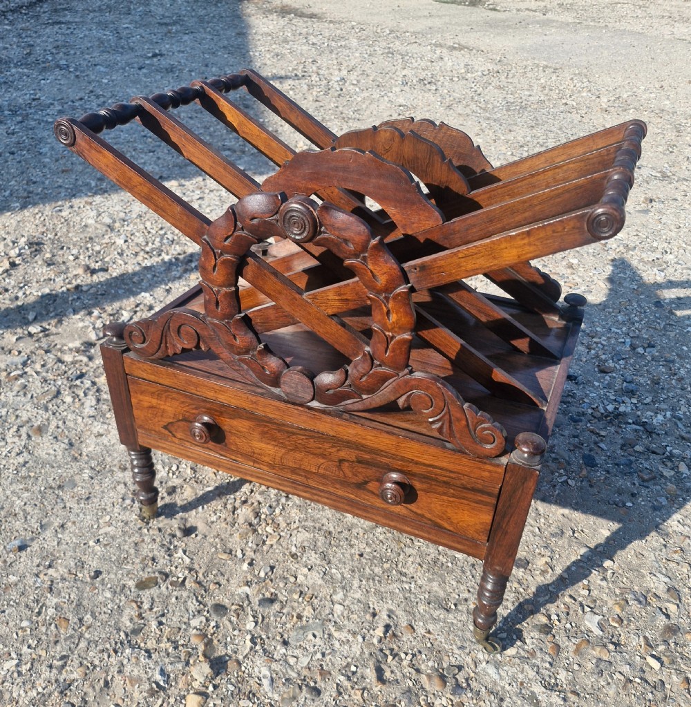 A Regency rosewood Canterbury featuring slatted division compartments, central carved leaf wreath motifs, a base drawer with turned handles, and turned legs on brass castors. View VI