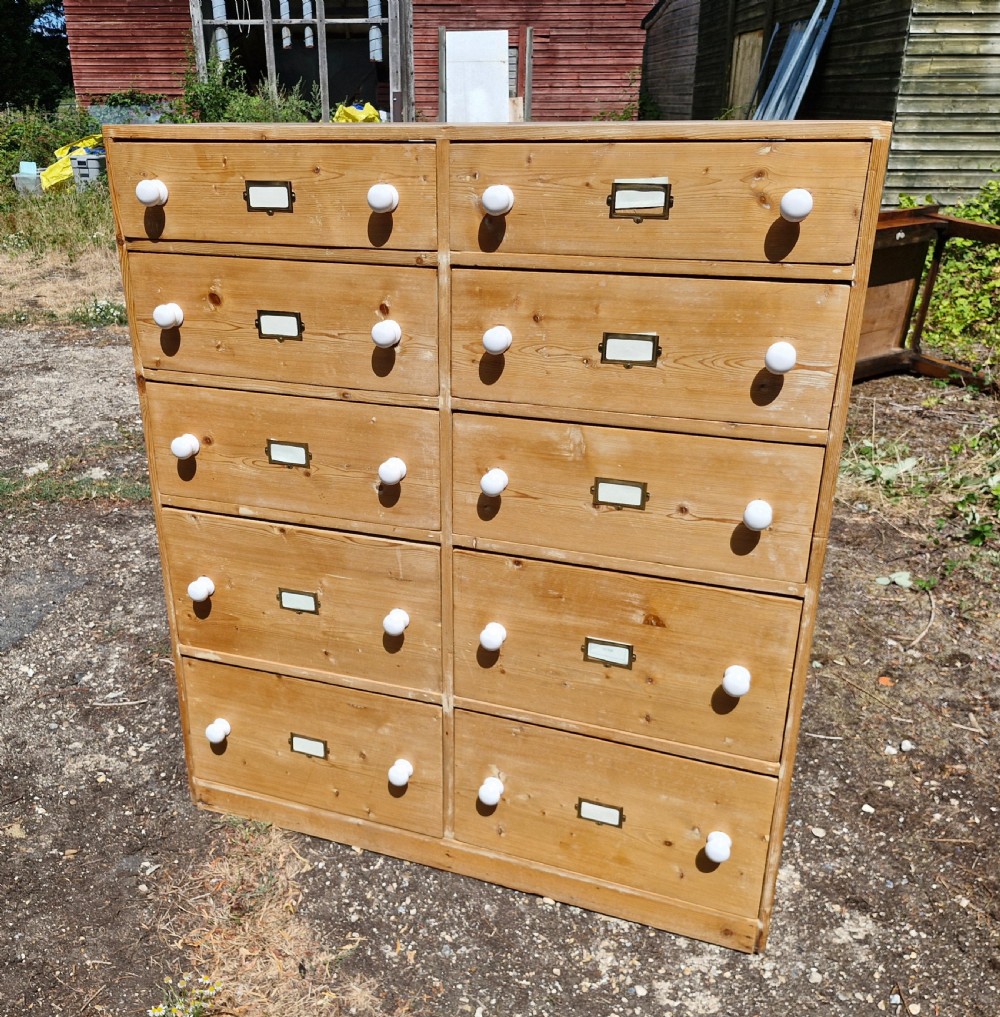 A large freestanding pine bank of drawers featuring ten drawers arranged in two columns with white ceramic knobs and brass label holders.