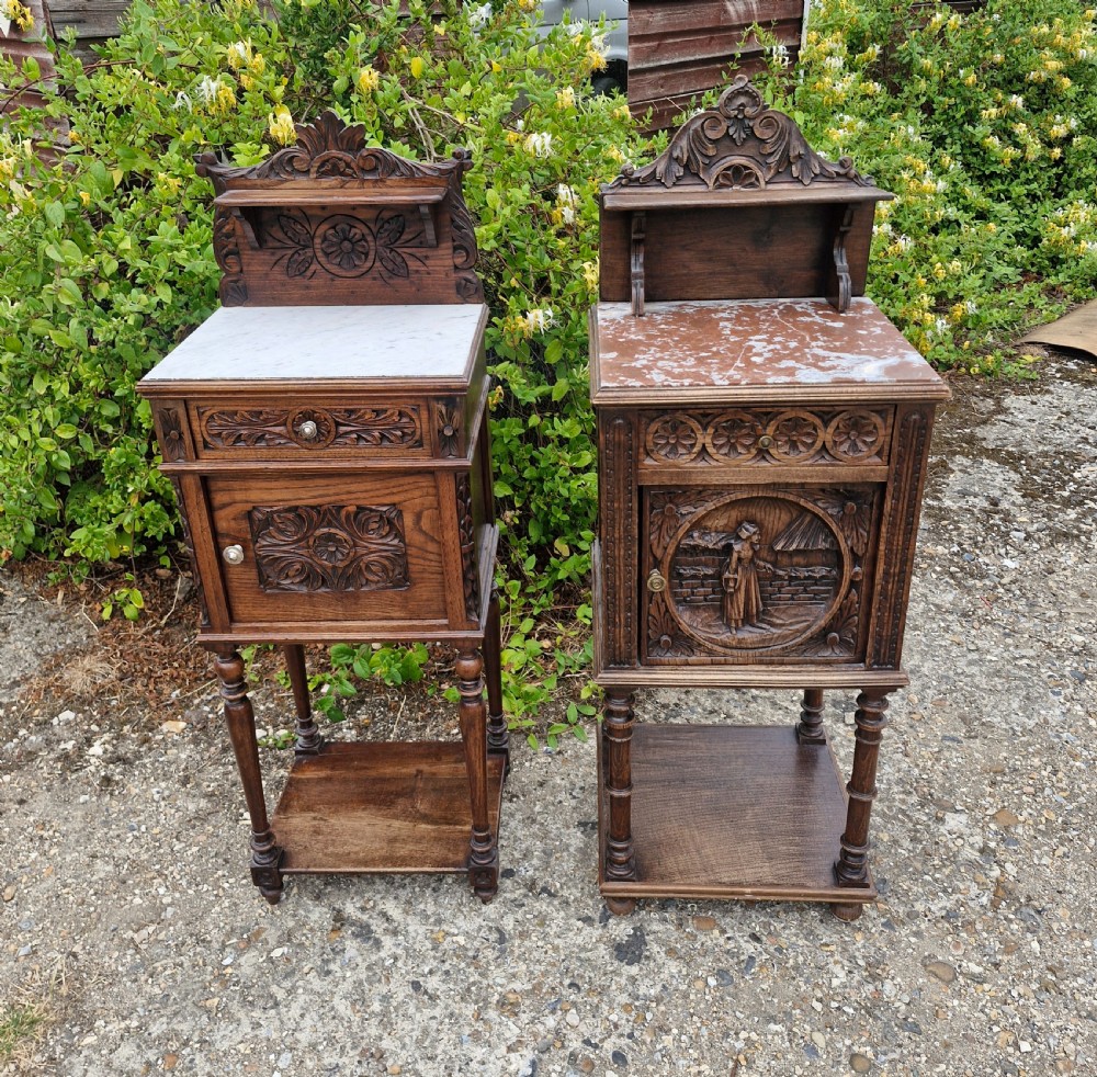 A pair of carved dark oak bedside cabinets featuring inset marble tops, relief panel cupboards, single drawers, and raised shelf cresting.