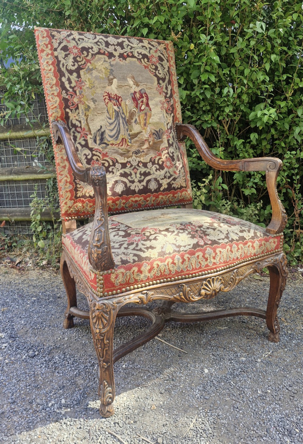 A late nineteenth-century high-back armchair featuring an intricately carved timber frame, scroll arms, cabriole legs, and pictorial needlework upholstery trimmed with brass studs.