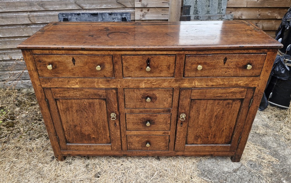 An early nineteenth-century Welsh oak cottage dresser featuring an open upper shelf rack above a base with multiple drawers and twin cupboard doors with brass handles. View VIII
