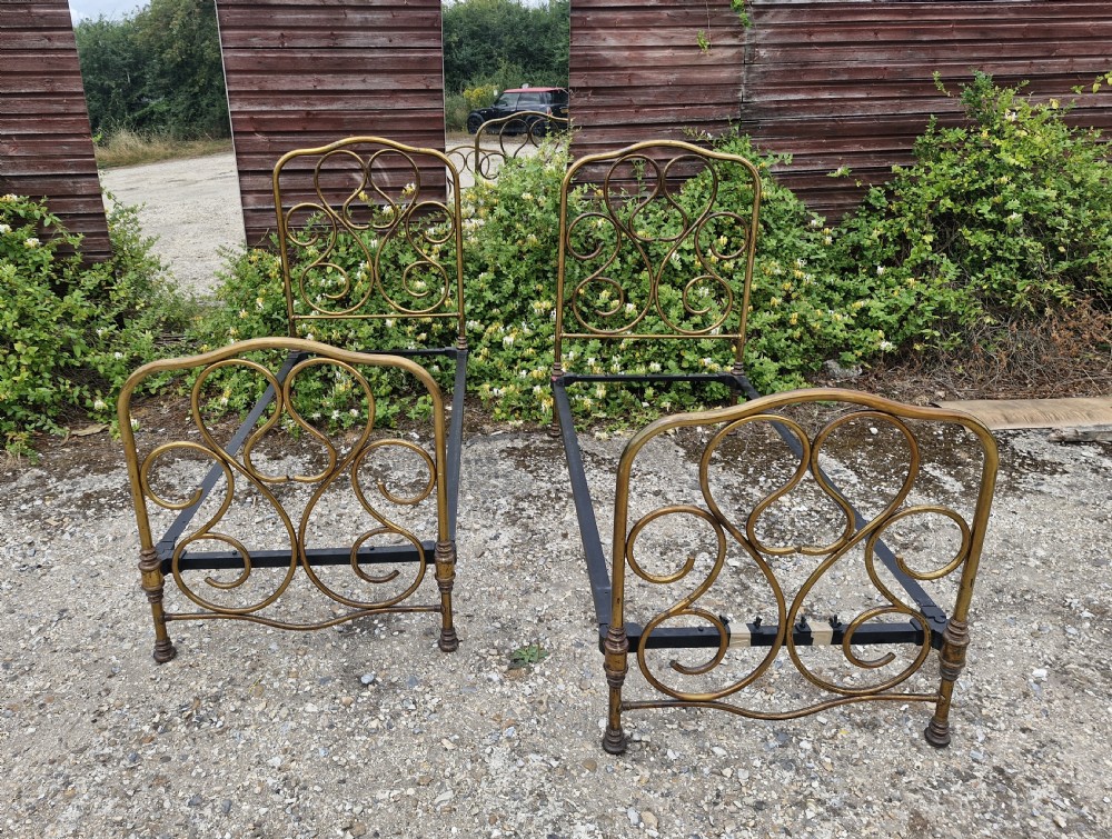 A matching pair of brass single bedsteads featuring decorative curved scrollwork headboards and footboards with black iron side rails. View IV
