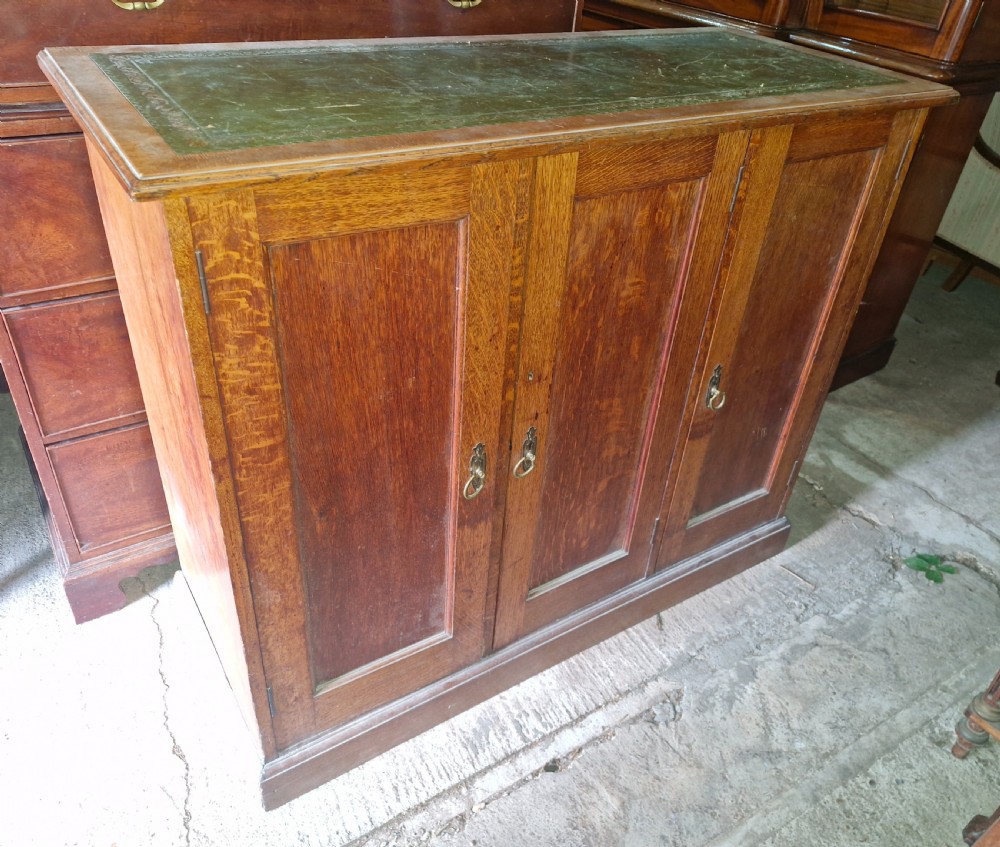 An antique golden oak cupboard featuring three panelled doors, brass ring handles, and an inset green leather top surface.